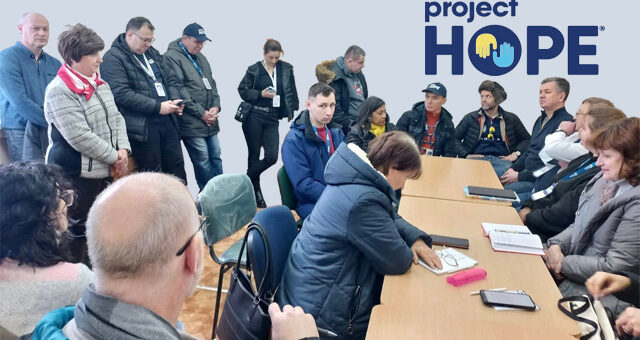 На зображенні група людей зібрана у кімнаті, кілька осіб сидять за столом, інші стоять поруч. Вони виглядають залученими в обговорення або нараду. У правому верхньому куті видно логотип "project HOPE". На столі розміщені блокноти, ручка, смартфон та рожевий циліндричний предмет. Зображення, ймовірно, пов'язане з колективним заходом чи проектом гуманітарного спрямування.