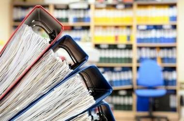 Ілюстрація: три великі різнокольорові папки з документами, на задньому фоні — стелаж з різнокольоровими  підписанимипапками