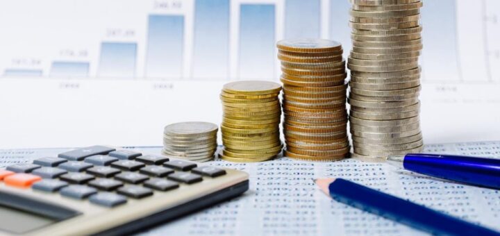 На столі лежить калькулятор, праворуч від нього - башенки з монет та олівець, на задньому фоні - діаграма