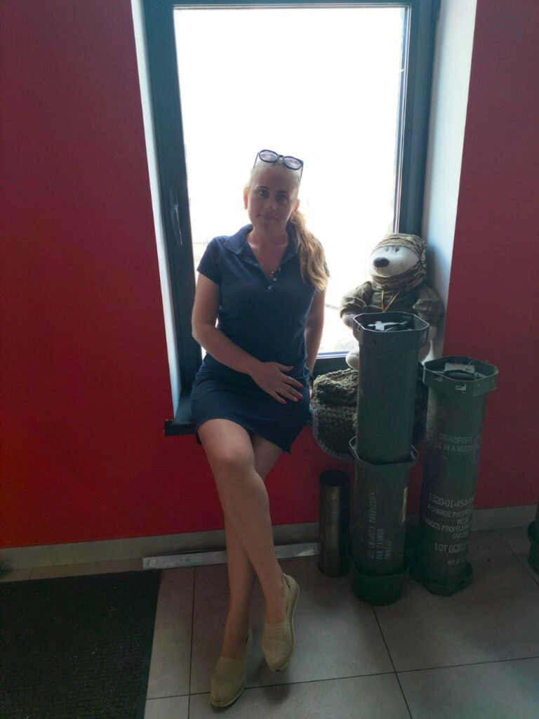 Співробітниця ХРД(В)А сидить на підвіконні, праворуч від неї частини від снарядів, а на них - іграшковий білий ведмедик в одязі ЗСУ та балаклаві