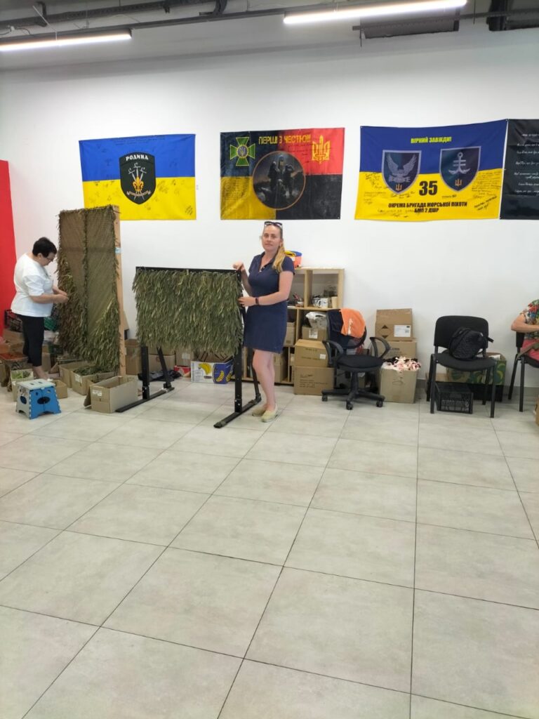 Співробітниця ХРД(В)А в залі поруч з готовою маскувальною сіткою для наших військових, позаду неї на стіні - прапори украйнських військових бригад