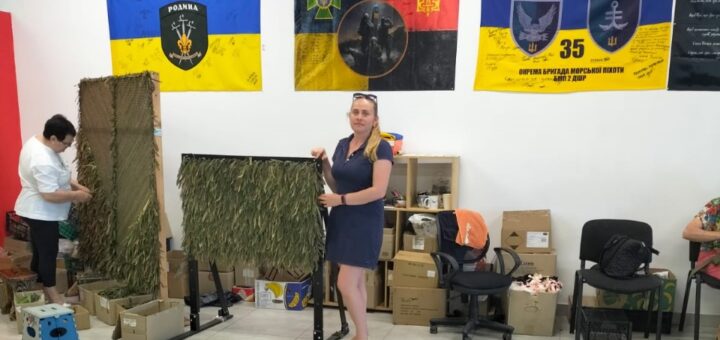 Співробітниця ХРД(В)А в залі поруч з готовою маскувальною сіткою для наших військових, позаду неї на стіні - прапори украйнських військових бригад