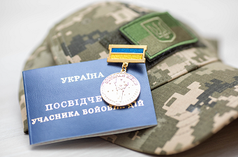 На зображенні — камуфляжна військова кепка з нашивкою у вигляді герба України, синя книжечка з написом українською «Україна. Посвідчення учасника бойових дій» та медаль, прикріплена до книжечки. Медаль має кольори українського прапора — синій і жовтий.