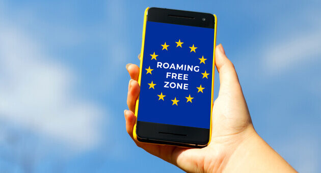 Смартфон в руці людини з написом на екрані 'ROAMING FREE ZONE' на фоні прапора Європейського Союзу. Фон – ясне блакитне небо.