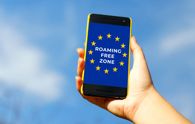 Смартфон в руці людини з написом на екрані 'ROAMING FREE ZONE' на фоні прапора Європейського Союзу. Фон – ясне блакитне небо.