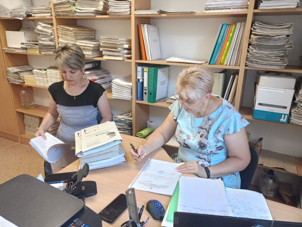 Дві жінки працюють в офісі, переглядаючи документи за столом. Навколо стелажі, заповнені паперами, папками та книгами.