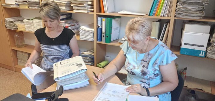 Дві жінки працюють в офісі, переглядаючи документи за столом. Навколо стелажі, заповнені паперами, папками та книгами.