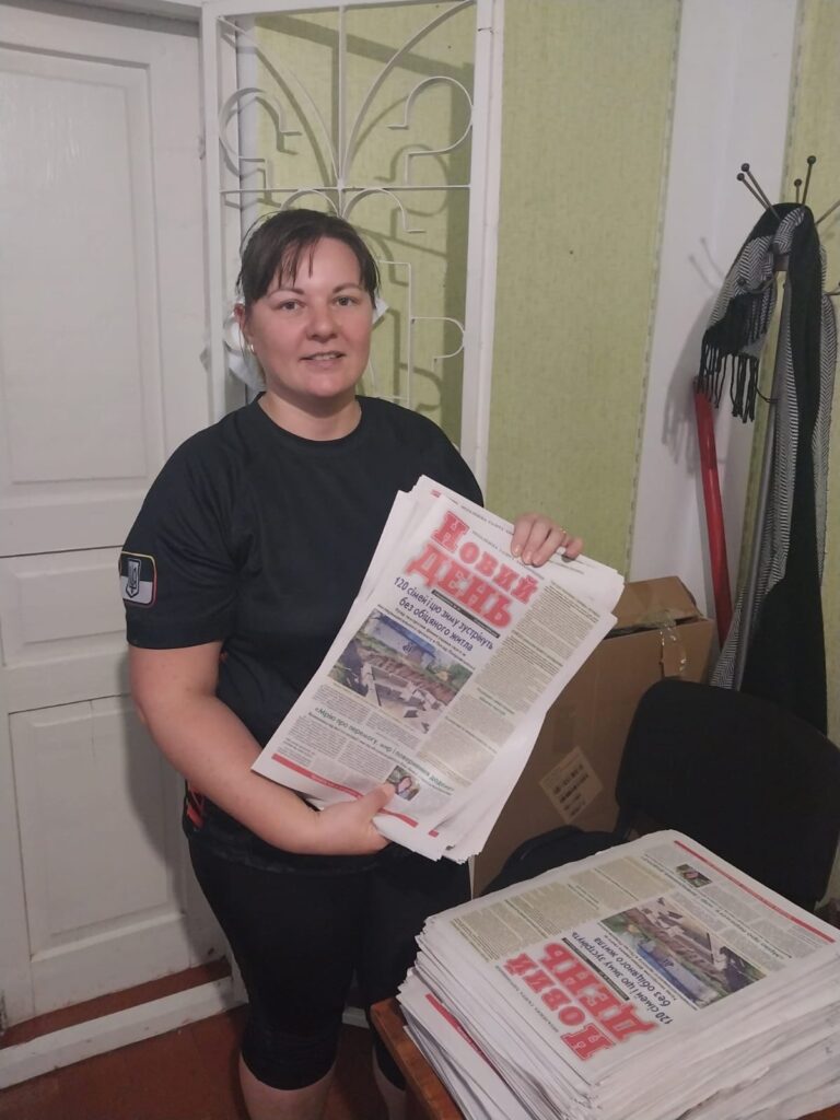 Дівчина у чорному одязі тримає газету "Новий день" та посміхається