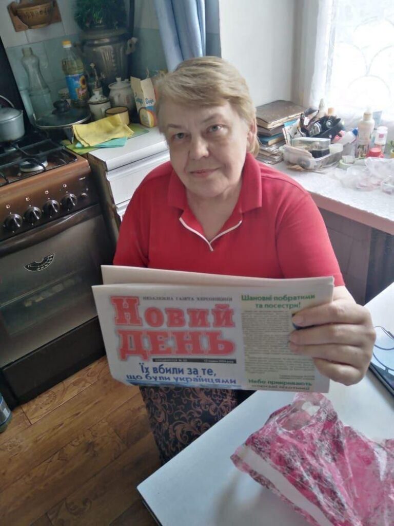 Жіночка у червоній сорочці на кухні сидить за столом та читає газету "Новий день"