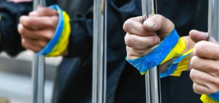 люди тримаються за ґрати, навколо їх рук обмотані жовто-блакитні стрічки