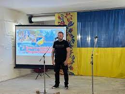 Дмитро Бутрій поздоровляє Чорнобаївку з днем народження, ліворуч від нього - плакат зі світлинами Чорнобаївки, праворуч - великий прапор України