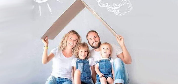 Всміхнена родина (тато, мама та двоє діточок) сидять на піблозі умовного будиночка, тримаючи його картонний дах