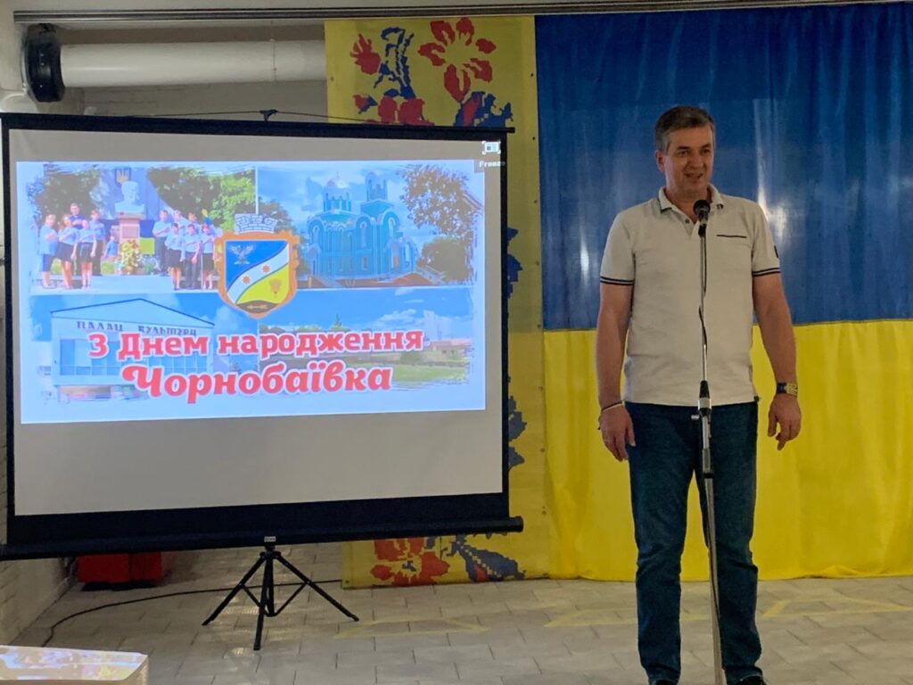 Михайло Валерійович Линецький поздоровляє Чорнобаївку з днем народження, ліворуч від нього - плакат зі світлинами Чорнобаївки, праворуч - великий прапор України