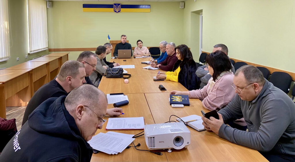 Начальники військових адміністрацій населених пунктів Херсонського району сидять у світлій кімнаті з гербом України за довгим столом, в голові столу — Михайло Линецький та Марина Малахова