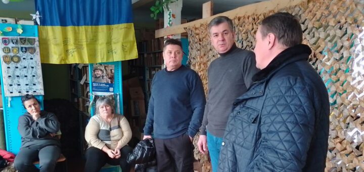 Михайло Линецький, Ігор Острівний та староста у приміщенні сільради. У кімнаті різні предмети, включаючи український прапор із підписами або повідомленнями, дисплей із значками або нашивками та камуфляжну сітку праворуч. Також є полиці з книгами та іншими речами на задньому плані. Підлога дерев'яна, а зі стелі звисає зелена лампа