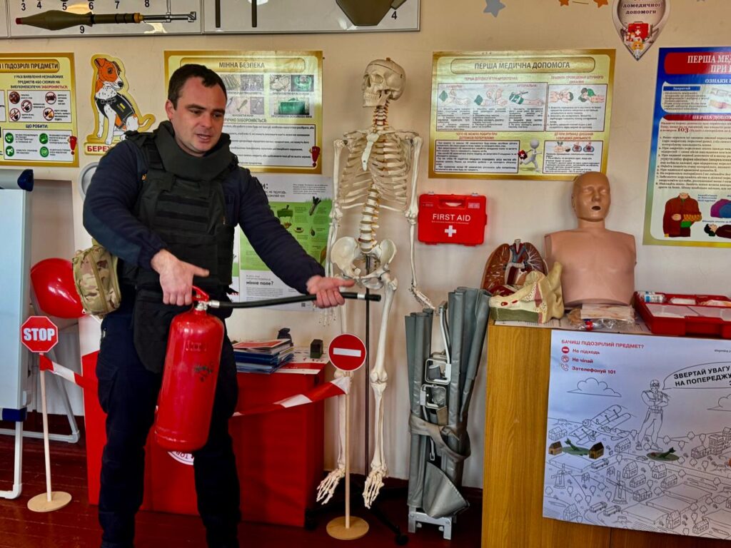 Рятувальник у класі пожежної безпеки показує вогнегасник, поруч з ним - різні плакати, манекени, тренажери тощо
