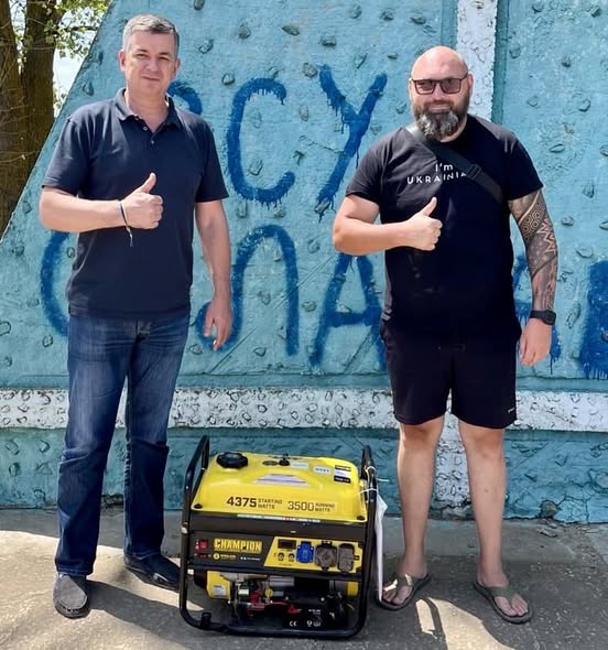 Михайло Линецький передає військовому жовтий генератор. Обидва показують жест "палець вгору". На генераторі зазначені характеристики: "4375 starting watts, 3500 running watts." На задньому плані видна синя стіна з написом ЗСУ СЛАВА!