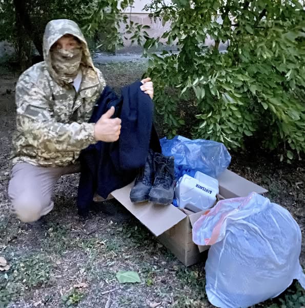 На фото людина у камуфляжній куртці з піднятим капюшоном, яка стоїть навколішки біля відкритої картонної коробки. Коробка містить різноманітні речі, включаючи чорні черевики, синій поліетиленовий пакет і білий контейнер із написом "KARCHER". Біля коробки розташований великий пластиковий пакет. Людина тримає в одній руці темний предмет одягу, а іншою рукою показує жест "палець угору". На задньому плані видно зелену рослинність