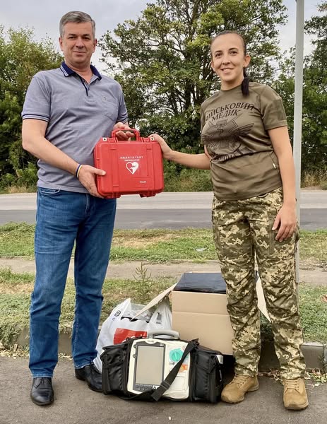 Михайло Линецький передає медичне спорядження жінці-військовослужбовцю у зеленій футболці та камуфляжних штанах. Вони разом тримають червоний кейс з білим символом серця. На землі перед ними видно чорну медичну сумку з відкритим обладнанням всередині, білий пластиковий пакет та картонну коробку. На задньому плані — дорога, зелень та дерева.