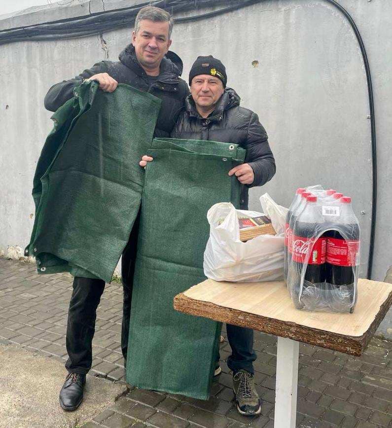 Михайло Линецький та боєць ЗСУ стоять на вулиці, тримаючи у руках маскувальні сітки. Обоє одягнені в темний одяг, включаючи куртки, і один з них у шапці-біні. Перед ними знаходиться маленький дерев'яний стіл, на якому лежить білий пластиковий пакет із деякими речами та упаковка з шести пляшок Coca-Cola. На задньому плані видно бетонну стіну з кабелями.