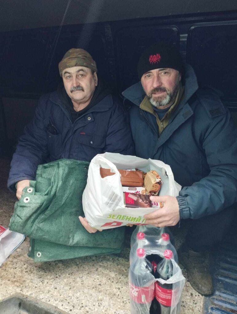 На фото двоє людей, які сидять у транспортному засобі і тримають у руках пакети з продуктами. Один з них тримає зелену маскувальну сітку, а інший — білий пластиковий пакет з різними продуктами.