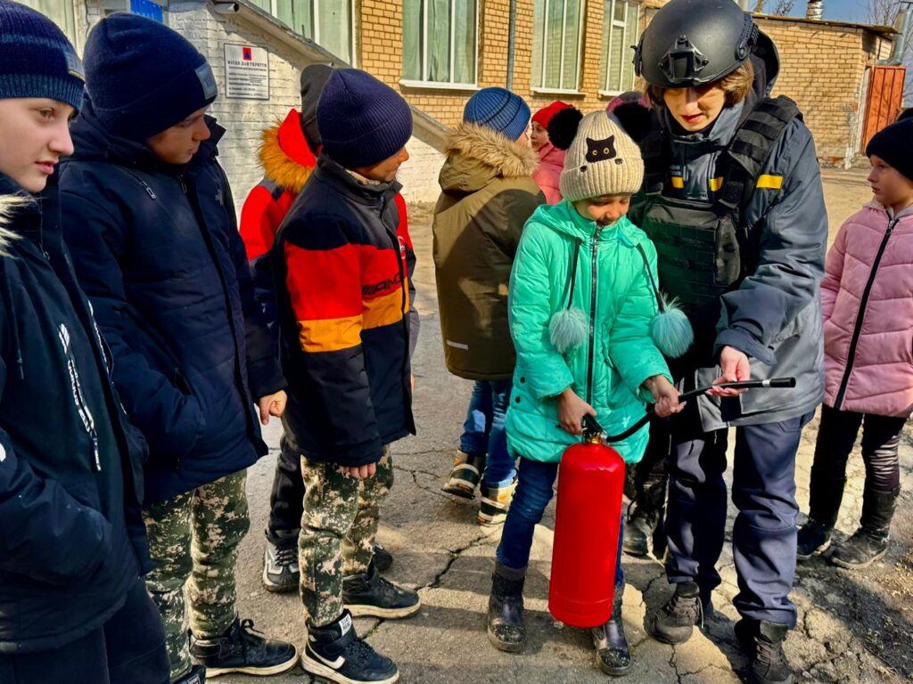 На фото зображено групу дітей та дорослого у захисному спорядженні, який проводить навчання з використання вогнегасника. Діти одягнені в зимовий одяг, стоять на вулиці біля будівлі з цегли. Один з дітей тримає червоний вогнегасник, а дорослий показує, як ним користуватися.