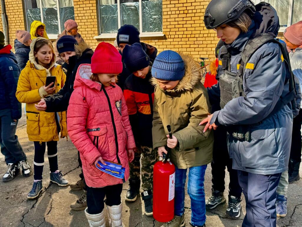 Діти стоять на вулиці біля будівлі з жовтої цегли. Діти одягнені в зимовий одяг, деякі з них тримають мобільні телефони. Один з дітей тримає вогнегасник, а поруч з ним стоїть людина в захисному одязі, яка проводить інструктаж або навчання з пожежної безпеки