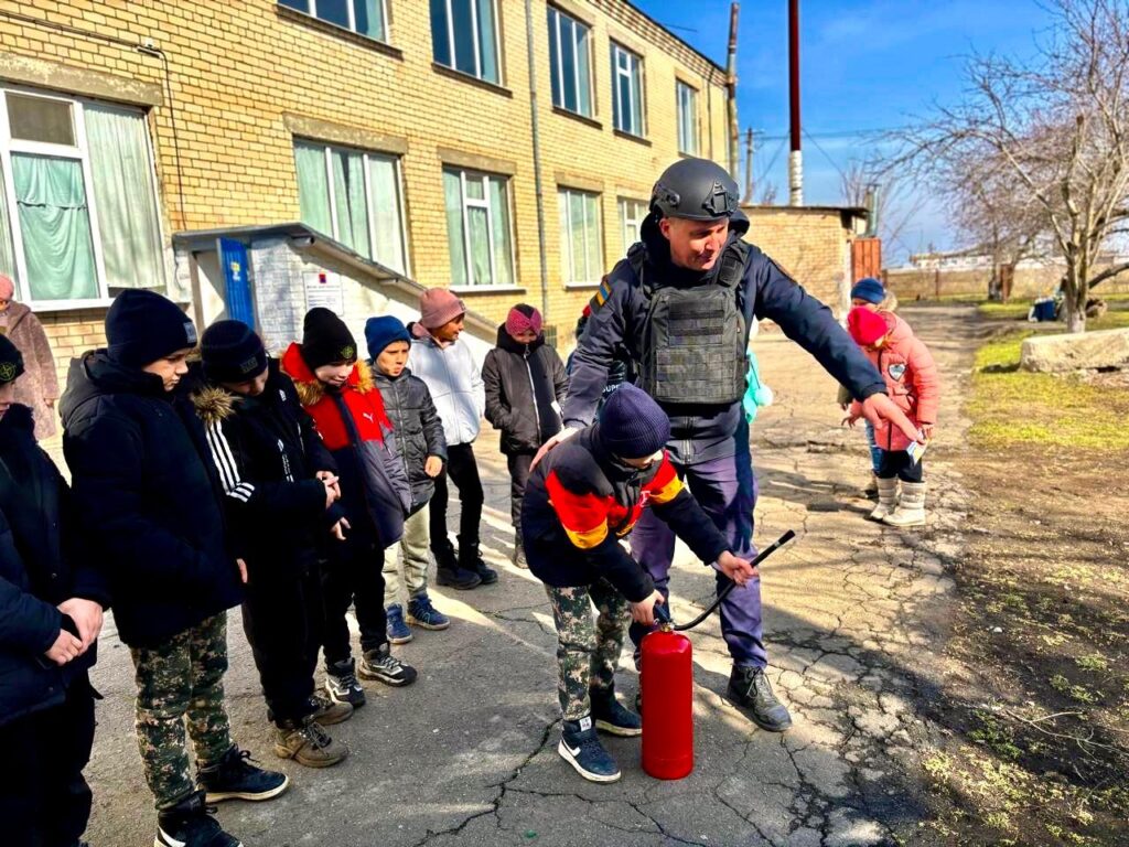 На фото зображено групу людей, які стоять на вулиці біля будівлі з жовтої цегли. Один з людей у захисному спорядженні допомагає іншій людині використовувати вогнегасник. Людина у захисному спорядженні вказує рукою на щось, а інша людина тримає вогнегасник і шланг. Інші люди стоять поруч і спостерігають за процесом. На задньому плані видно дерева та інші будівлі.