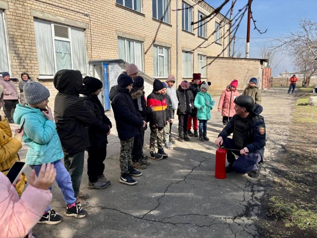 На фото зображено групу дітей, які стоять на вулиці перед цегляною будівлею з кількома вікнами. Вони стоять у лінію і дивляться на дорослого в формі та шоломі, який демонструє використання вогнегасника. Діти одягнені у зимовий одяг