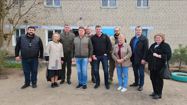 На фото зображена група з дев'яти людей, які стоять на вулиці перед будівлею, зведеною з білої цегли. Люди одягнені в куртки, пальта та інший повсякденний одяг. На задньому плані видно металеві ґрати на вікнах будівлі та сині козирки над ними. На передньому плані розташовані зелені насадження й автомобільні шини, пофарбовані в зелений колір, які використовуються як клумби.