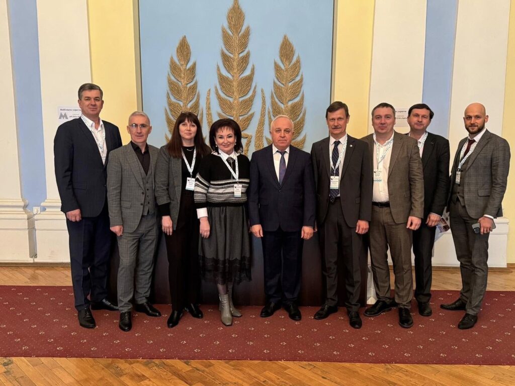 Вісім людей на фоні декоративної стіни із зображенням трьох великих колосків пшениці. Всі присутні одягнуті у формальний одяг: чоловіки в костюмах, а жінки у сукнях. На шиї в деяких із них є бейджі або іменні значки, що свідчить про участь у конференції чи офіційному заході. Приміщення має дерев’яну підлогу та червону доріжку.