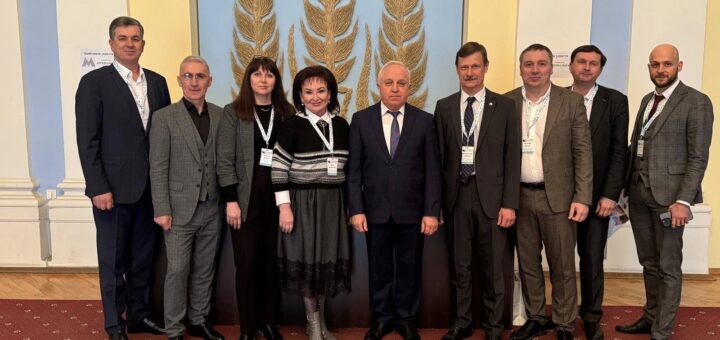 Вісім людей на фоні декоративної стіни із зображенням трьох великих колосків пшениці. Всі присутні одягнуті у формальний одяг: чоловіки в костюмах, а жінки у сукнях. На шиї в деяких із них є бейджі або іменні значки, що свідчить про участь у конференції чи офіційному заході. Приміщення має дерев’яну підлогу та червону доріжку.