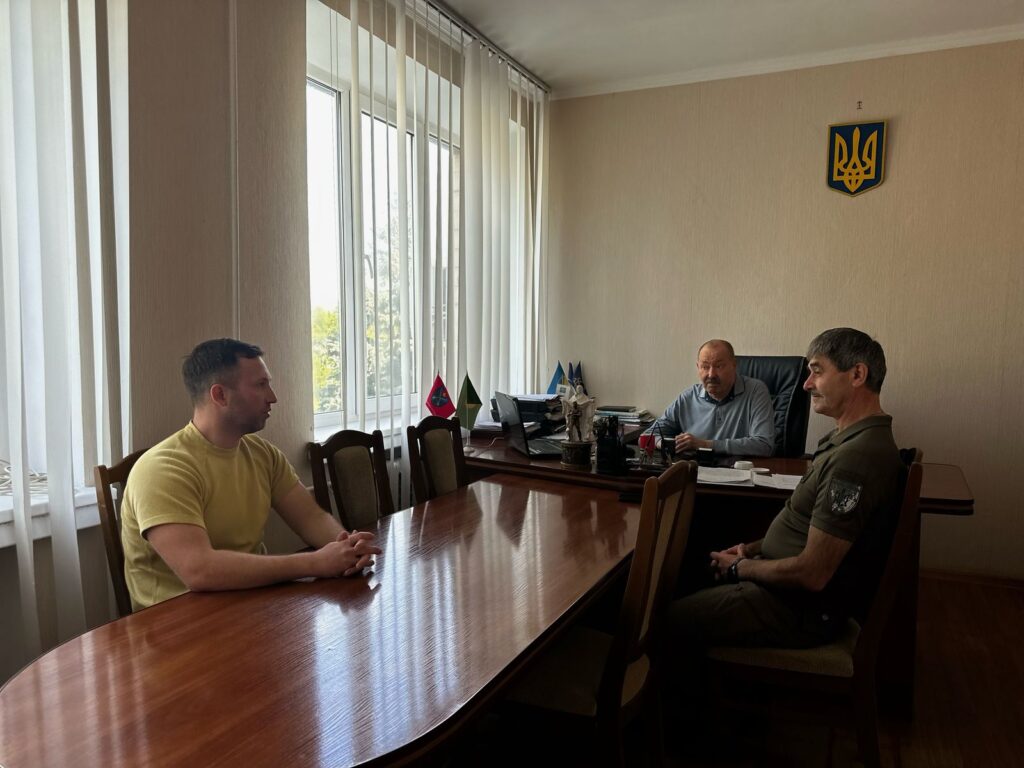 На фото зображено трьох осіб, які сидять за дерев'яним столом у офісному просторі. Позаду видніються великі вікна з вертикальними жалюзі, які пропускають природне світло. На стіні за столом розташований герб України. На столі знаходяться різні предмети, серед яких комп'ютер, статуетка та кілька прапорів. Серед прапорів є український прапор та прапор із червоно-синім дизайном. 