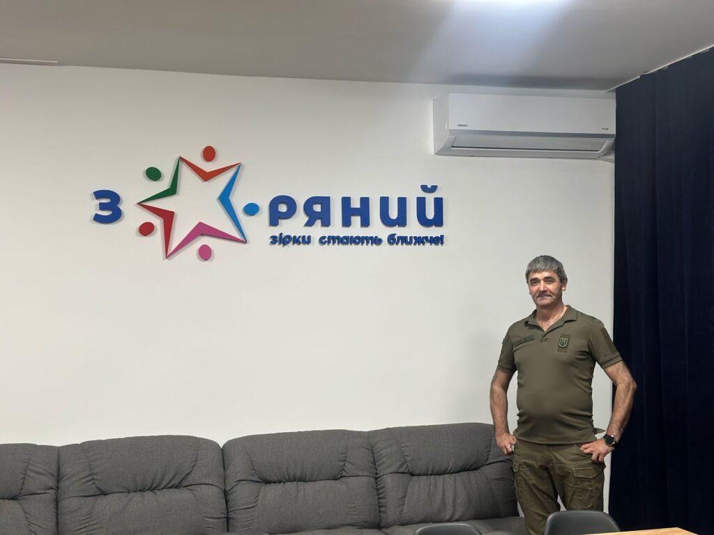 На фото зображено Олександра Бабуцького у військовій формі. Він стоїть біля стіни, на якій розміщено логотип та напис. Логотип складається з п'ятикутної зірки з різнокольоровими кінцями (зелений, червоний, синій, рожевий) та крапками навколо неї. Поруч із логотипом написано "Зоряний" та "зірки стають ближчі". Олександр стоїть біля сірого дивана, а на стіні видно кондиціонер.