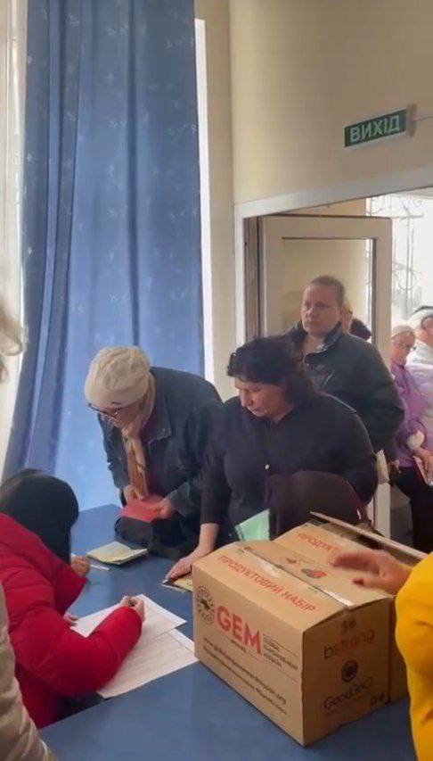 На знімку група людей стоїть у черзі всередині кімнати. Справа за столом сидить людина в червоній куртці, яка пише щось на папері. На столі велика картонна коробка з написом "GEM" та "Good Neighbors". На коробці є текст іншою мовою. У кімнаті сині штори, а над дверима вивіска з написом «ВИХІД», що означає «exit» українською