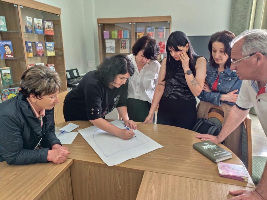 На зображенні група людей, які сидять навколо столу в приміщенні, що нагадує бібліотеку або кімнату для навчання. Один із них працює над великим аркушем паперу, який лежить на столі, малюючи чи пишучи щось. Інші спостерігають за процесом. На задньому плані видно стелажі, заповнені книжками.