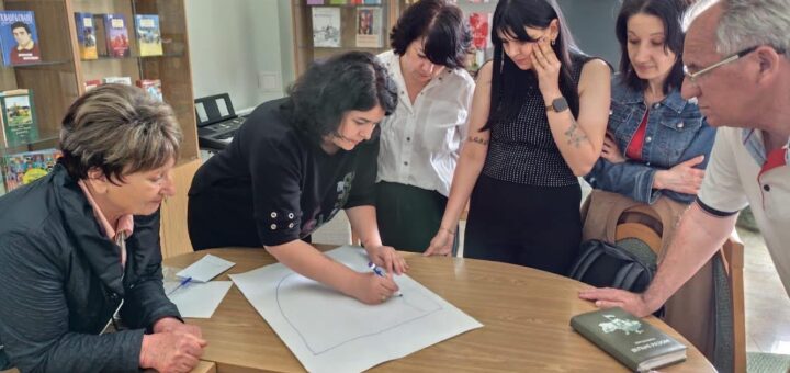 На зображенні група людей, які сидять навколо столу в приміщенні, що нагадує бібліотеку або кімнату для навчання. Один із них працює над великим аркушем паперу, який лежить на столі, малюючи чи пишучи щось. Інші спостерігають за процесом. На задньому плані видно стелажі, заповнені книжками.