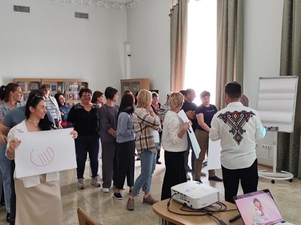 На зображенні група людей у кімнаті з книжковими стелажами на задньому плані. У центрі зображення учасник тримає великий аркуш паперу, на якому намальований червоний півколо та чотири вертикальні лінії всередині. Біля нього інші люди сидять чи стоять, спостерігаючи за процесом. На столі попереду розташований проектор і ноутбук із зображенням людини на екрані. Праворуч стоїть фліпчарт із чистим папером.