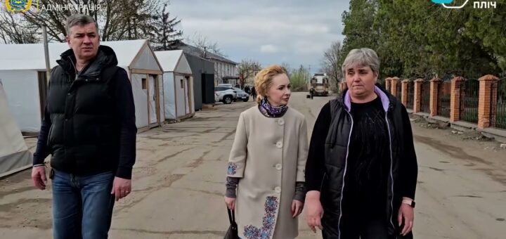 Михайло Линецький, Мар’яна Томин і Лариса Соколова. йдуть вулицею Чорнобаївської громади. Вони рухаються поруч із наметами та будівлями. На задньому плані видно автомобілі та дерева.