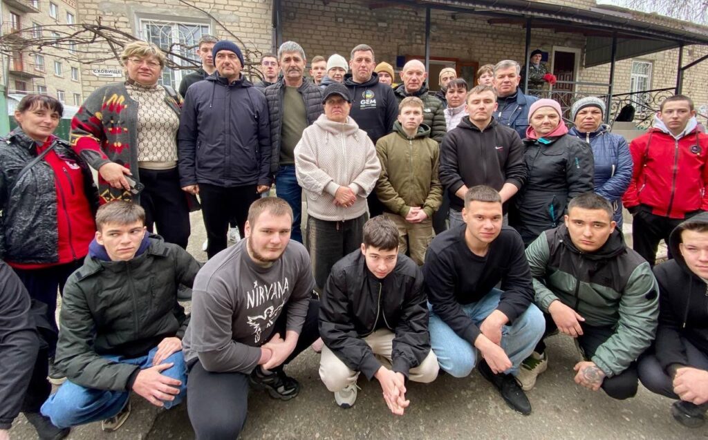 На знімку група приблизно з двадцяти людей стоїть та сидить на колінах перед будівлею. Люди одягнені в різний повсякденний одяг, серед якого є куртки, светри та худі. Деякі з них носять шапки або кепки. На задньому плані видно будівлю з вікнами, дверима та написом «СХОВИЩЕ».