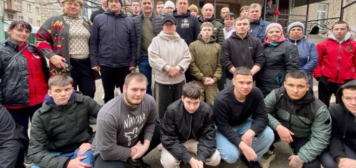 На знімку група приблизно з двадцяти людей стоїть та сидить на колінах перед будівлею. Люди одягнені в різний повсякденний одяг, серед якого є куртки, светри та худі. Деякі з них носять шапки або кепки. На задньому плані видно будівлю з вікнами, дверима та написом «СХОВИЩЕ».