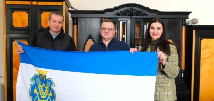 На фото три людини тримають прапор Херсонської області, Україна. Прапор має три горизонтальні смуги: верхня і нижня синього кольору, середня – білого. У центрі білої смуги знаходиться герб Херсонської області, що складається із жовтої корони, синього щита із зображенням жовтої пшениці та інших символів. На задньому плані видно меблі з дерев’яними елементами та чорними й помаранчевими деталями.