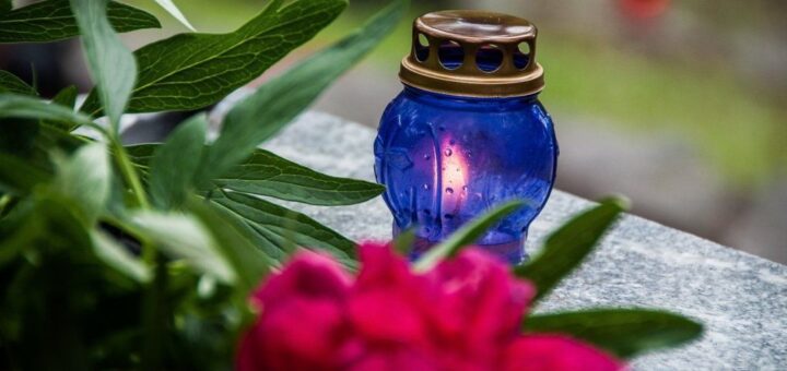 На зображенні свічка у синьому скляному ліхтарі з металевою кришкою. Кришка бронзового кольору, має круглі отвори. Навколо ліхтаря лежать зелене листя та червона квітка, схожа на піон. Фон розмитий