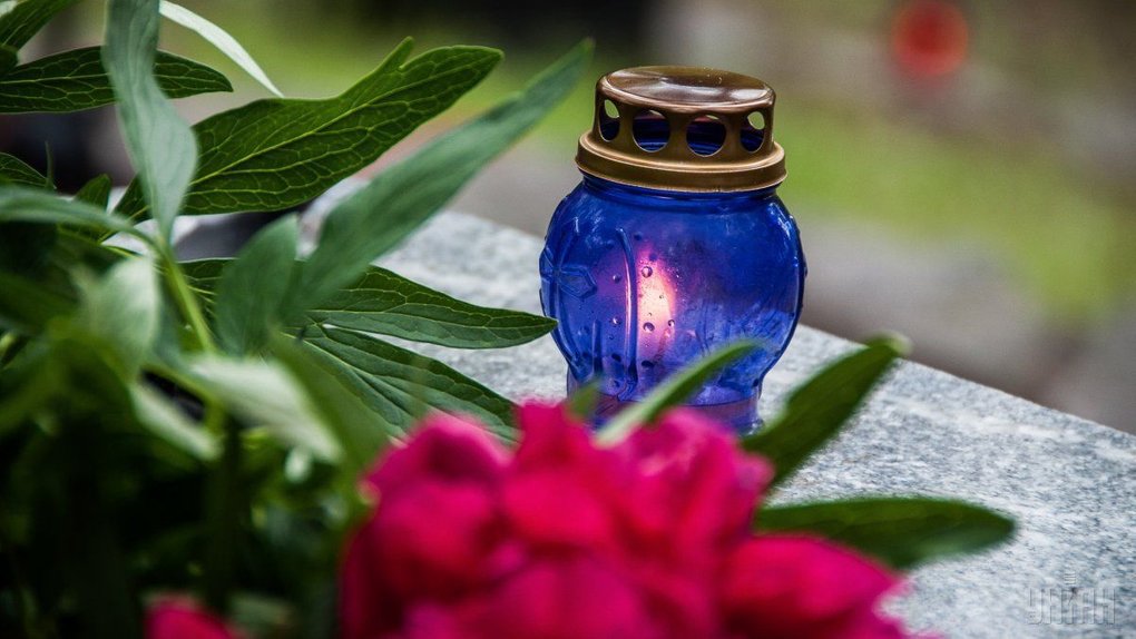 На зображенні свічка у синьому скляному ліхтарі з металевою кришкою. Кришка бронзового кольору, має круглі отвори. Навколо ліхтаря лежать зелене листя та червона квітка, схожа на піон. Фон розмитий