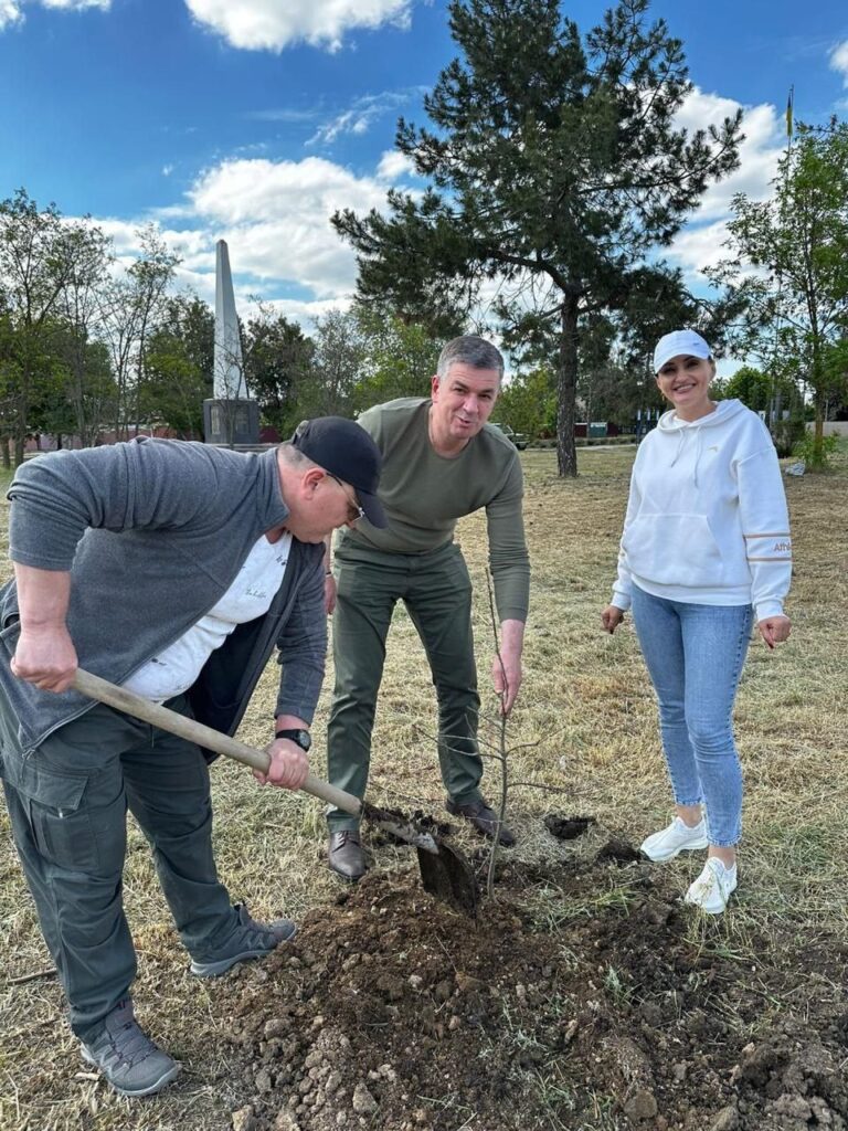 На зображенні видно три особи, які займаються висадкою дерева на відкритій місцевості. Одна людина використовує лопату для викопування ями, друга утримує саджанець, готуючи його до посадки, а третя уважно спостерігає за процесом. На задньому плані видно зелені газони, кілька дерев та монумент, що створюють атмосферу громадської або екологічної активності. Небо напівхмарне, підкреслюючи природний характер сцени.