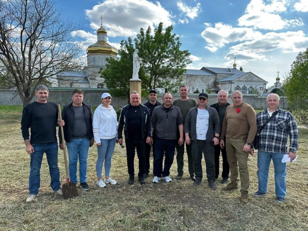 На зображенні показано групу людей, що стоять на зеленій трав'янистій галявині перед релігійною спорудою з золотими куполами, на яких розташовані хрести – це, напевно, церква чи собор. Один з учасників, що знаходиться серед групи, тримає лопату, що може свідчити про участь у певній заході або діяльності на відкритому повітрі. За групою розташована статуя на підмостках, а на задньому плані видніються дерева. Небо частково покрито хмарами з ділянками блакитного кольору, що створює природну атмосферу.