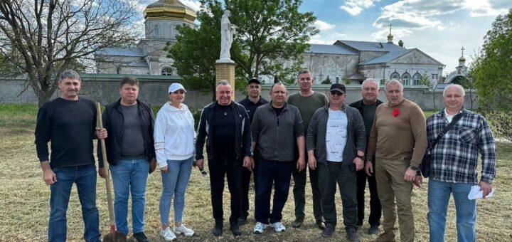 На зображенні показано групу людей, що стоять на зеленій трав'янистій галявині перед релігійною спорудою з золотими куполами, на яких розташовані хрести – це, напевно, церква чи собор. Один з учасників, що знаходиться серед групи, тримає лопату, що може свідчити про участь у певній заході або діяльності на відкритому повітрі. За групою розташована статуя на підмостках, а на задньому плані видніються дерева. Небо частково покрито хмарами з ділянками блакитного кольору, що створює природну атмосферу.