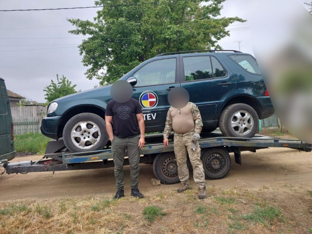 На фотографії двоє людей стоять перед темно-зеленим позашляховиком, що завантажений на платформний причіп. На дверях водія автомобіля є круглий емблемний знак із багатоколірним дизайном і написом «TEV» під ним. Люди стоять на ґрунтовій дорозі, обабіч якої росте трава. Позаду видно дерев’яну огорожу та дерева. Автомобіль виглядає як офіційний або військовий транспортний засіб, враховуючи одяг одного з присутніх.
