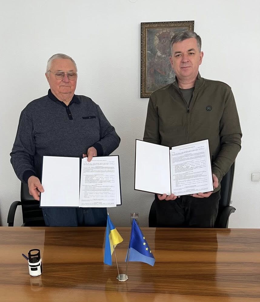 Попов Віктор Григорович та Линецький Михайло Валерійович підписали Меморандум про співпрацю. Вони стоять за дерев'яним столом, кожен тримає документ у руках. На столі розташовані два невеликі прапори: один представляє Україну, а інший — Європейський Союз. Також на столі є синій циліндричний об'єкт та ручка. Кімната має світлі стіни, а на одній з них висить картина у рамці. Це офіційний момент підписання угоди, що підтверджує співпрацю між сторонами.