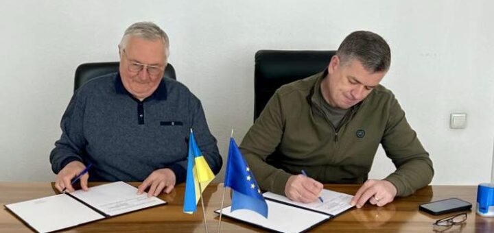 Попов Віктор Григорович та Линецький Михайло Валерійович підписують Меморандум про співпрацю. Вони сидять за дерев'яним столом у конференц-залі. На столі розташовані український та європейський прапор, документи для підпису, смартфон та синій циліндричний предмет. Кімната має світло-зелені стіни. На одній із них висить картина у рамці. Освітлення забезпечують стельові лампи, які рівномірно освітлюють простір. Обстановка підкреслює офіційний характер зустрічі та важливість укладання угоди.